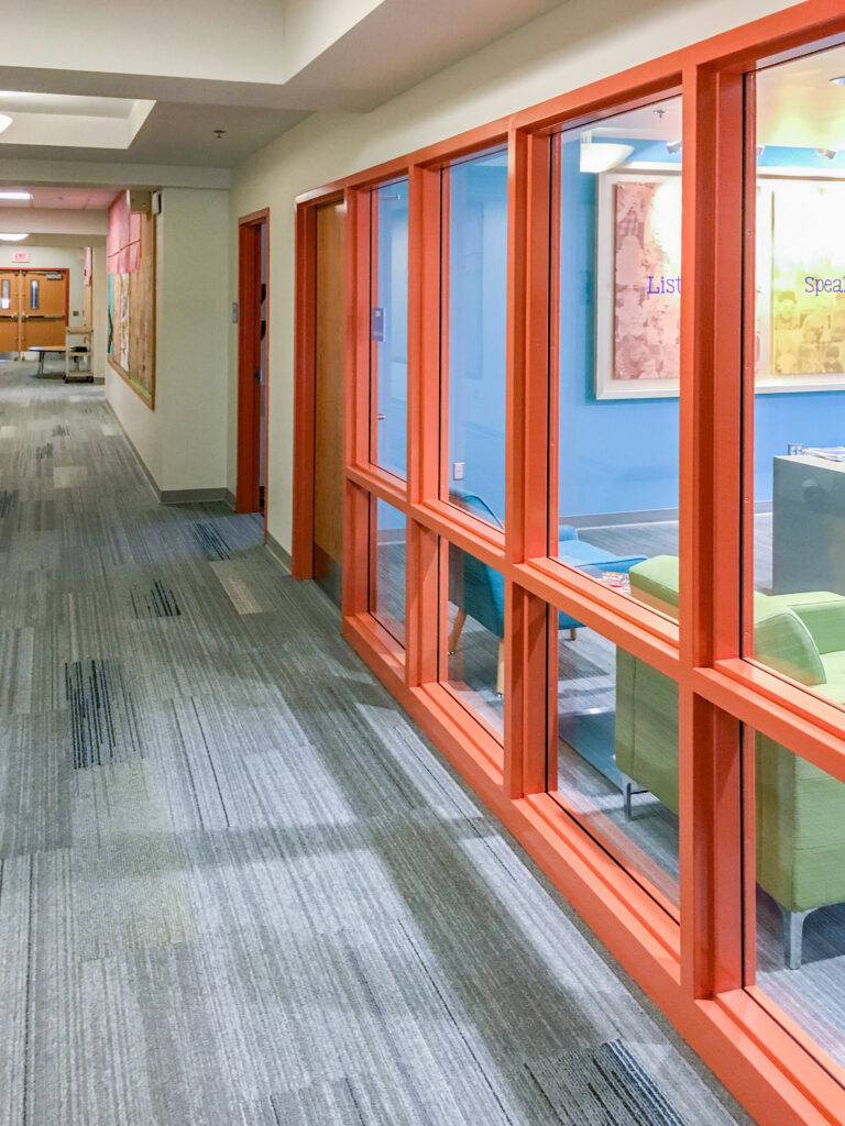A long office hallway with gray carpet and bright orange window frames looking into a blue-walled room with lounge chairs.