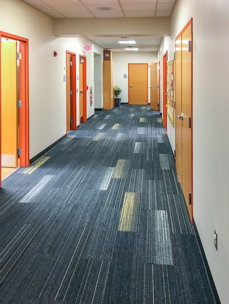 A long hallway lined with multiple orange and wood-toned doors, featuring gray-patterned carpet and an exit sign overhead.