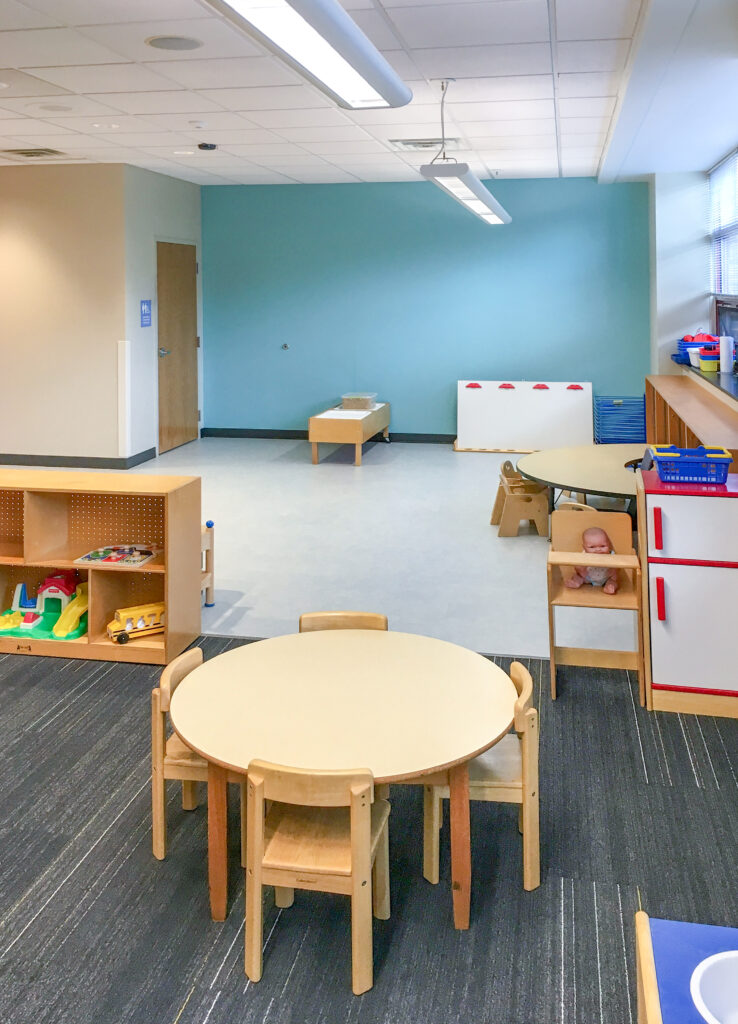 Preschool classroom with circular tables, small wooden chairs, open storage shelves, and a toy kitchen.