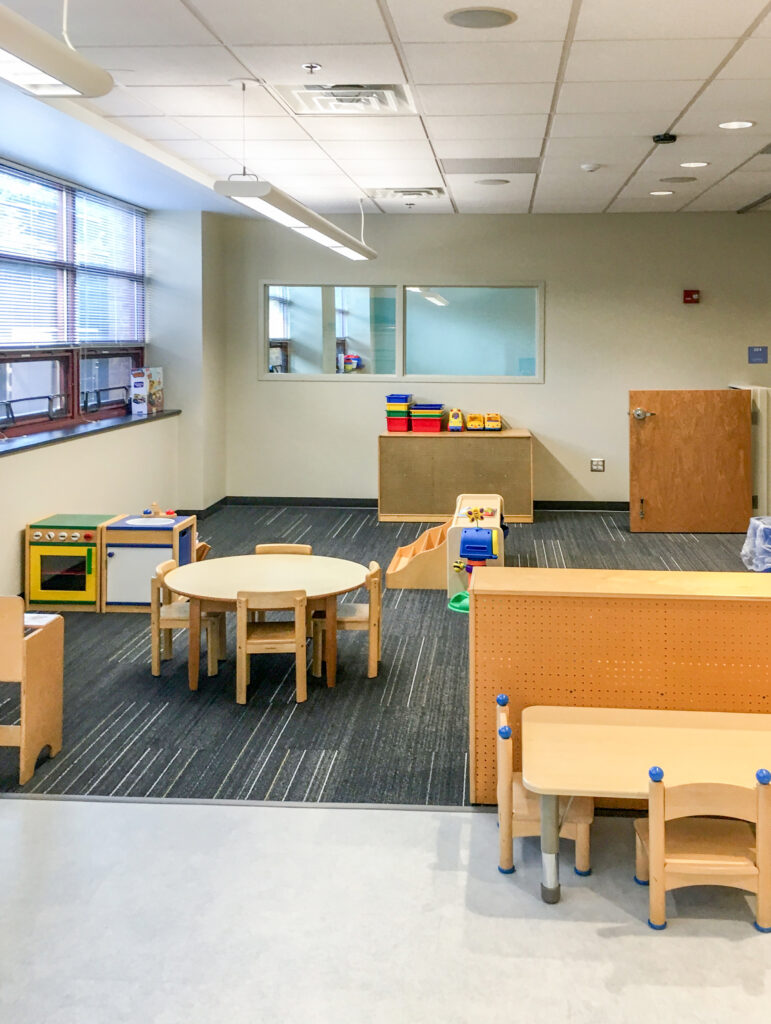 A brightly lit preschool classroom with small wooden tables, chairs, a play kitchen, and storage shelves.