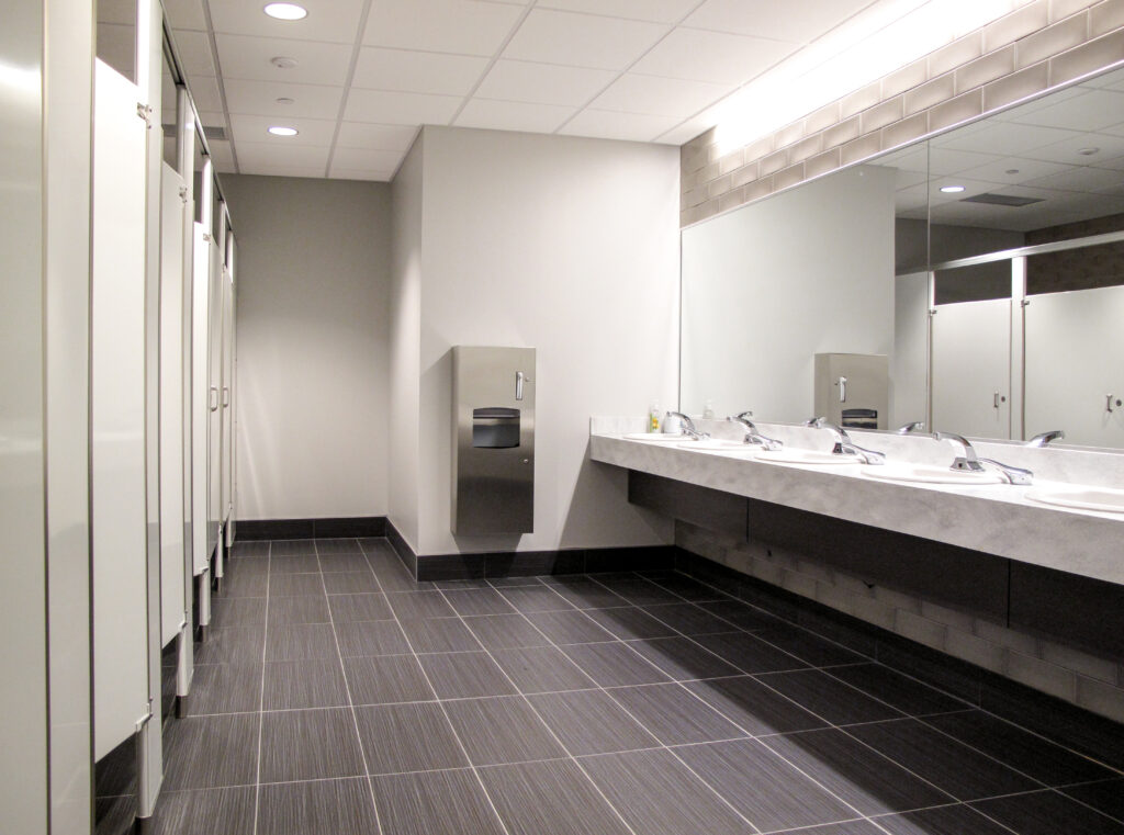 Public restroom with a row of toilet stalls, a wall-mounted paper towel dispenser, and a long vanity with multiple sinks and a large mirror.