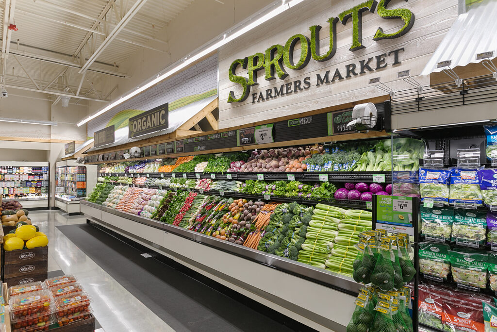 Produce section of a Sprouts Farmers Market store with shelves stocked with fresh vegetables, including carrots, broccoli, celery, and bagged salads.