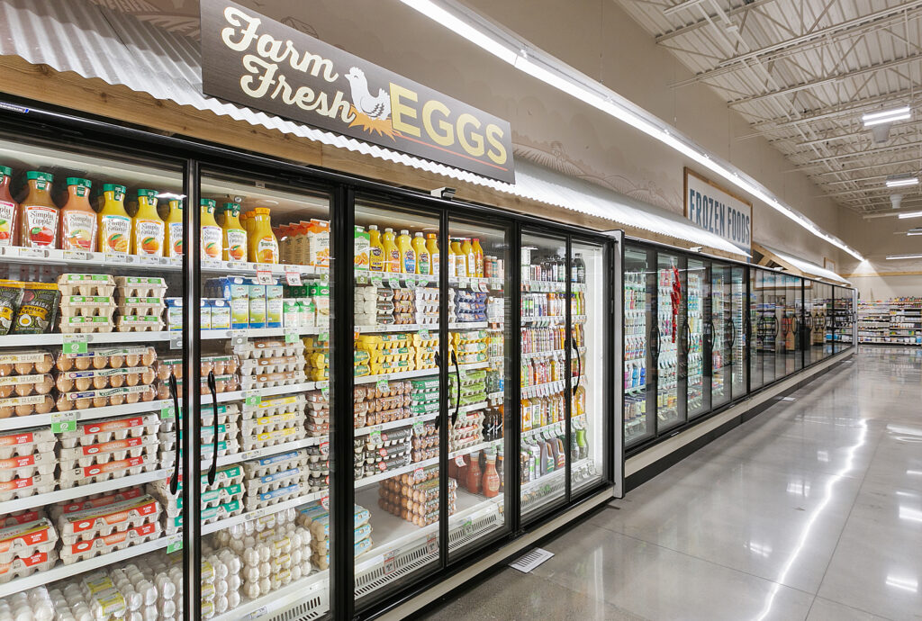 Grocery store refrigerated aisle featuring shelves stocked with cartons of eggs and orange juice, with signage reading “Farm Fresh Eggs.”