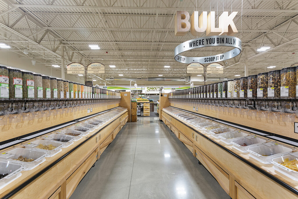 A grocery store aisle featuring two long rows of bulk food dispensers and open bins for self-service items, with overhead signs labeling categories like nuts, grains, and candy.