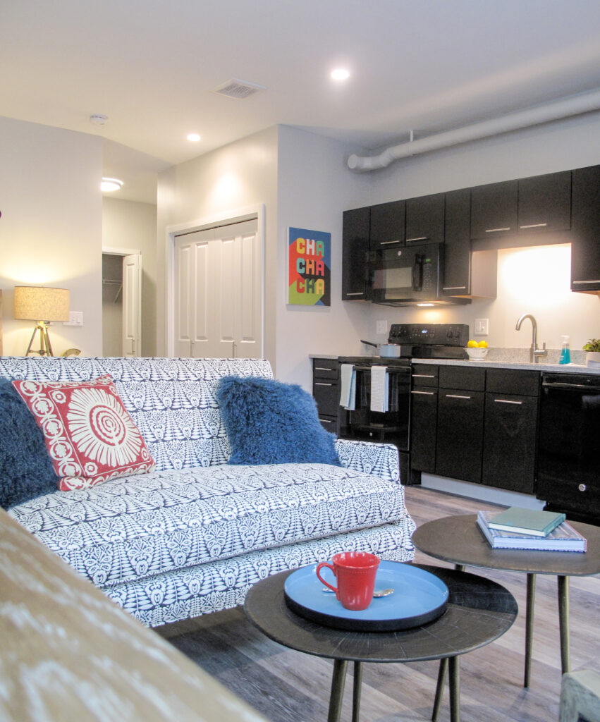 A patterned sofa with blue and red pillows faces two small round coffee tables in a living area connected to a kitchen with black cabinets.