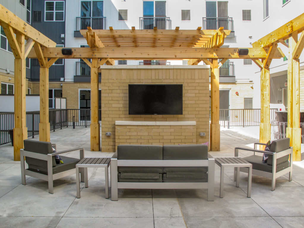 Outdoor lounge area with a mounted flat-screen TV on a brick wall under a wooden pergola, featuring a sofa and two chairs.