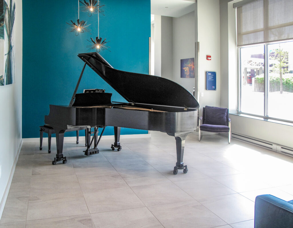 A black grand piano stands in a bright room with a teal accent wall and large windows.
