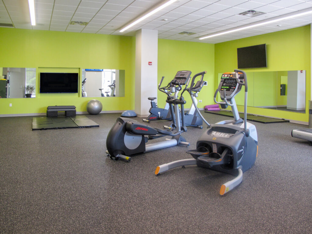 A gym with two elliptical machines in the foreground, exercise mats, an exercise ball, and mirrors on lime green walls.