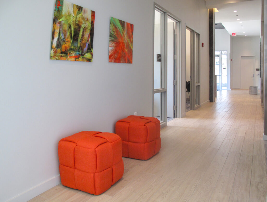 Two orange cube ottomans placed against a hallway wall under two abstract paintings, with glass-fronted office doors visible down the corridor.