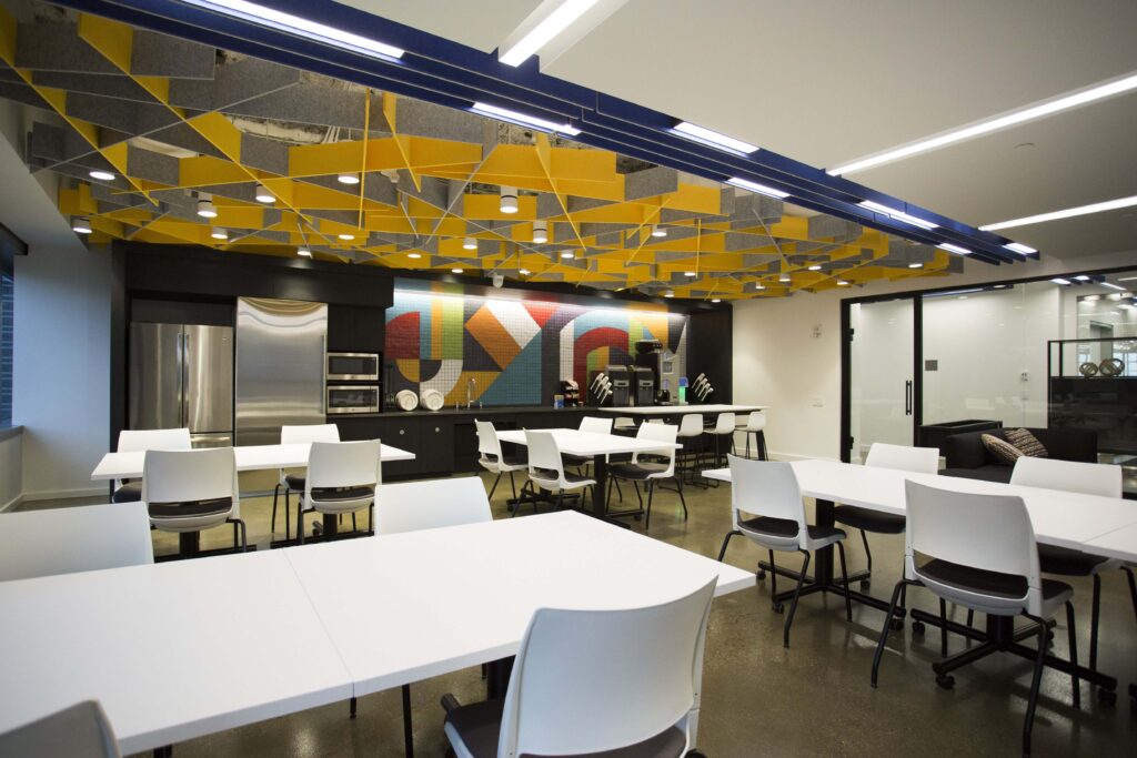 An office breakroom with several white tables, chairs, and a kitchen area featuring a colorful geometric tiled backsplash and a yellow suspended ceiling structure.