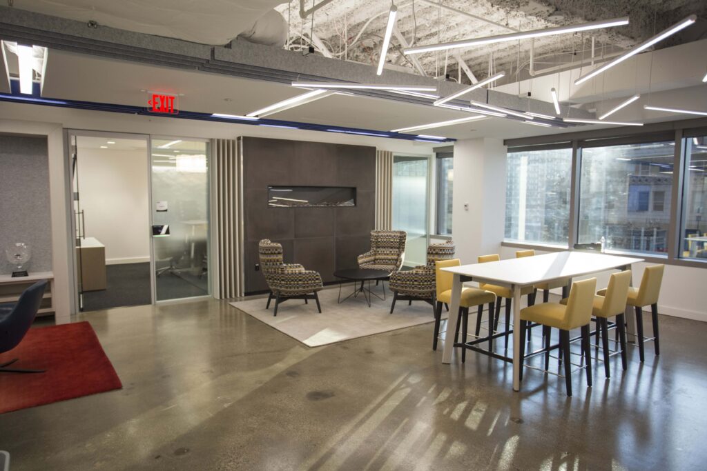 A modern office lounge with a seating area featuring three patterned chairs, a small table, a linear fireplace, and a high-top table with six yellow chairs.
