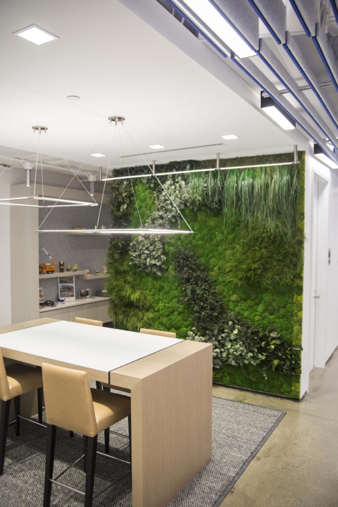 A wooden high-top table and chairs sit in front of a floor-to-ceiling interior wall covered in various live plants and moss.