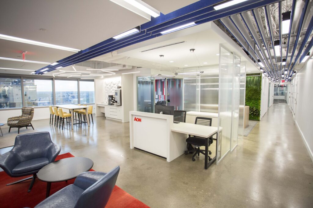 An Aon office reception area with a white desk, a seating area with blue chairs on a red rug, and glass-walled workspaces.