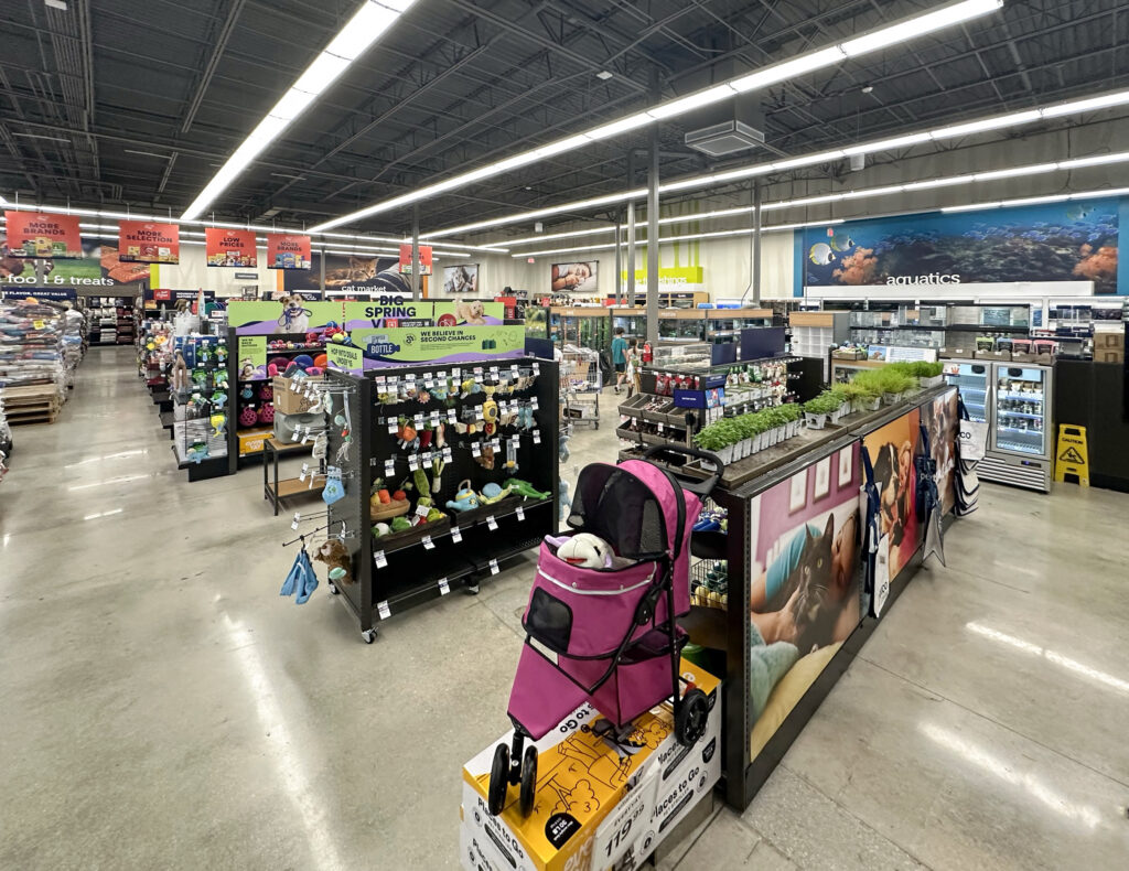 Interior of a pet supply store with aisles of toys, accessories, and an aquatics department in the background, featuring a pink pet stroller in the foreground.