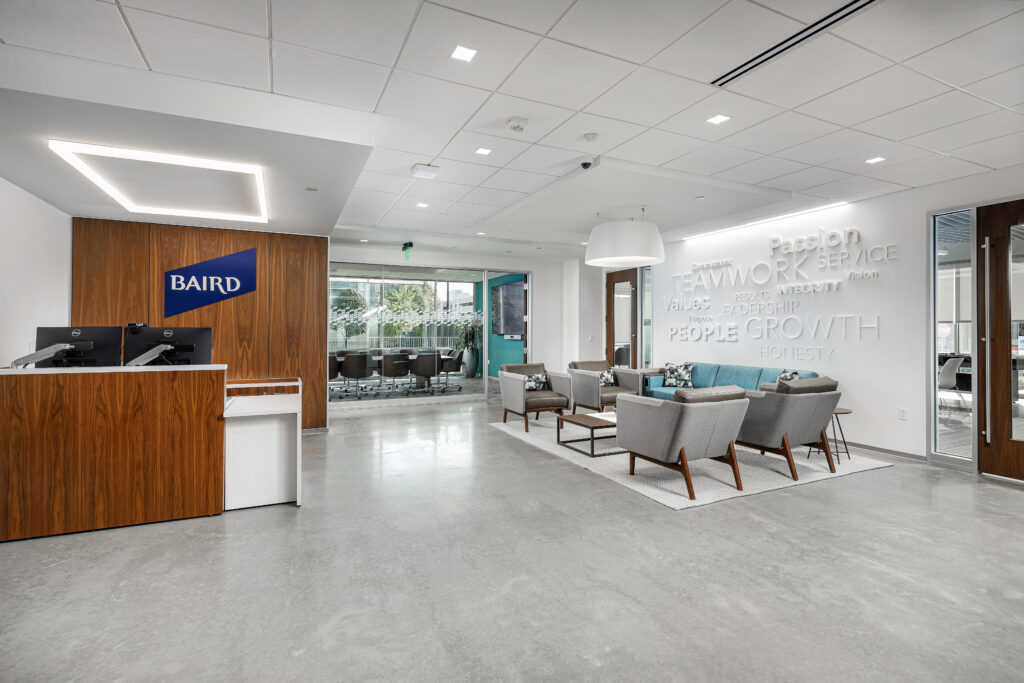 A modern office reception area featuring a Baird branded desk and a lounge space with armchairs, a sofa, and wall-mounted lettering displaying company values.