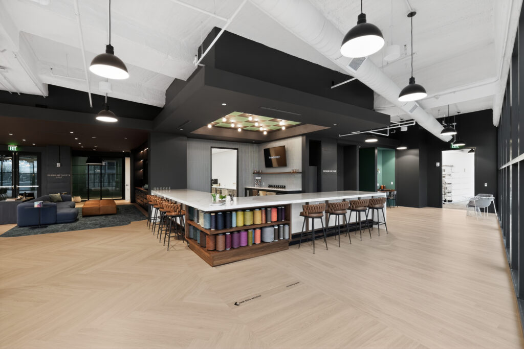 A modern office break room with a white L-shaped bar counter, brown stools, and shelves displaying colorful spools of thread.