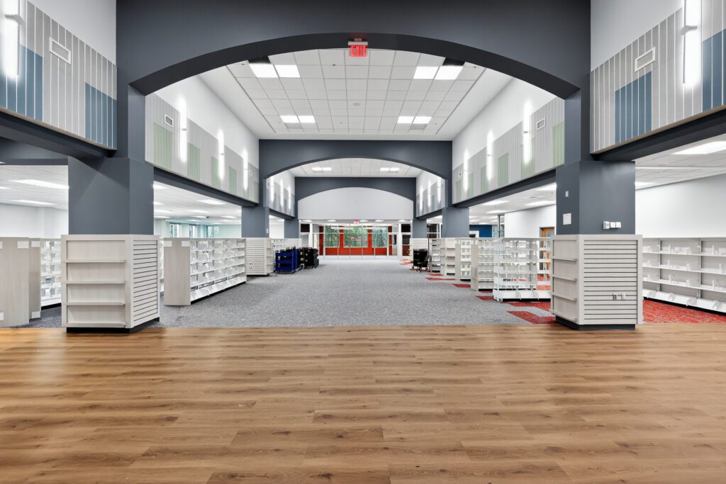 A wide, open view of a library interior featuring wooden floors in the foreground, rows of empty bookshelves, and a central walkway leading toward red exterior doors.