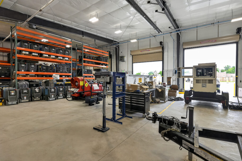 Interior of a warehouse featuring storage racks with stacked containers, a portable heater, a workbench with tools, and a trailer-mounted generator near an open bay door.
