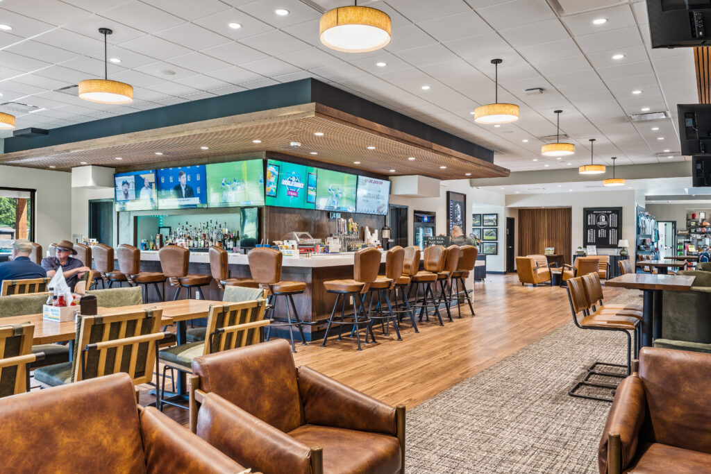 A spacious bar and restaurant lounge with leather armchairs, wooden tables, and a row of stools at a long bar featuring large screens displaying sports.