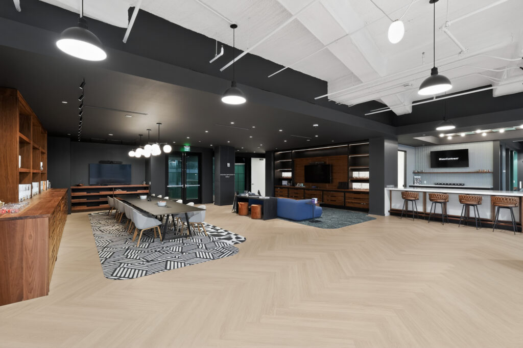 A modern office lounge featuring a long conference table, a blue sofa area, a bar with stools, and a large wood-paneled wall unit with an integrated television.