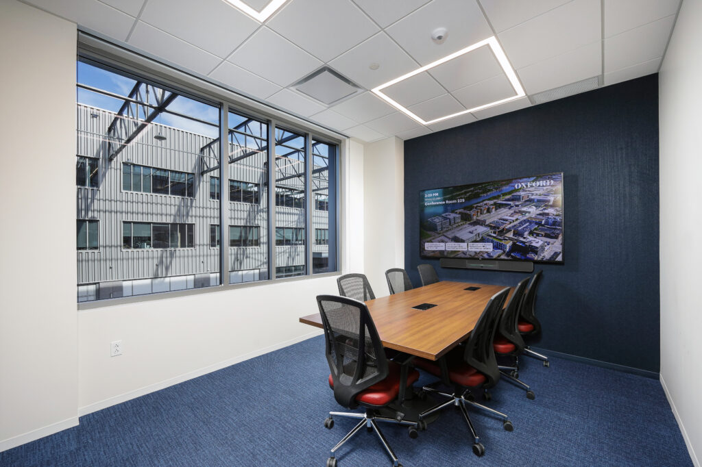 A conference room with a wooden table, six rolling chairs, and a wall-mounted display screen showing a map of the Oxford campus.