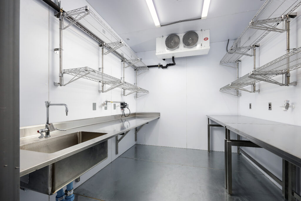 Interior of a commercial kitchen space with stainless steel counters, a sink, wall-mounted wire shelving, and a ceiling-mounted refrigeration unit.