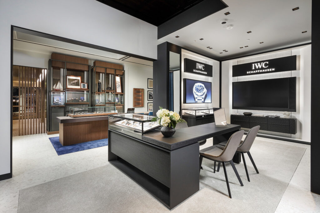 Interior of an IWC Schaffhausen boutique featuring display cases, a desk with two chairs, and wall-mounted screens with brand signage.