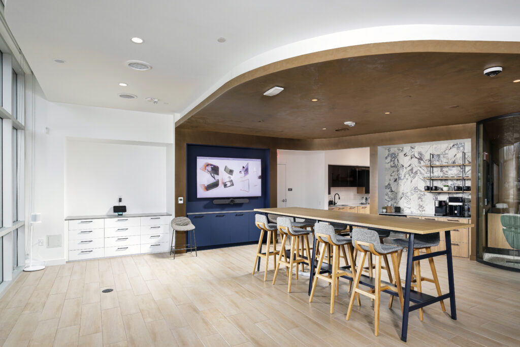 Modern office break room with a long wooden high-top table, six upholstered stools, and a wall-mounted television.