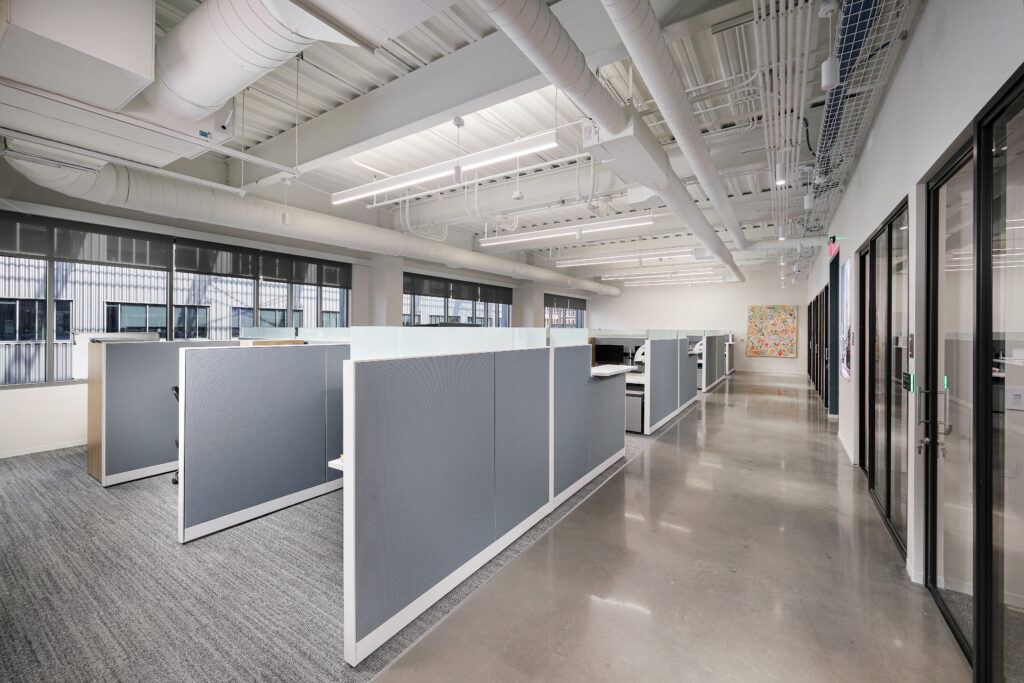 An open-plan office space featuring rows of grey cubicles, a polished concrete floor, and exposed ceiling ductwork.