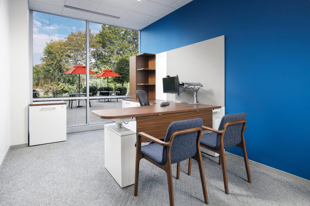 A modern office desk with a computer monitor, two blue chairs, and a tall wooden shelf, positioned in front of a large window overlooking a patio with red umbrellas.