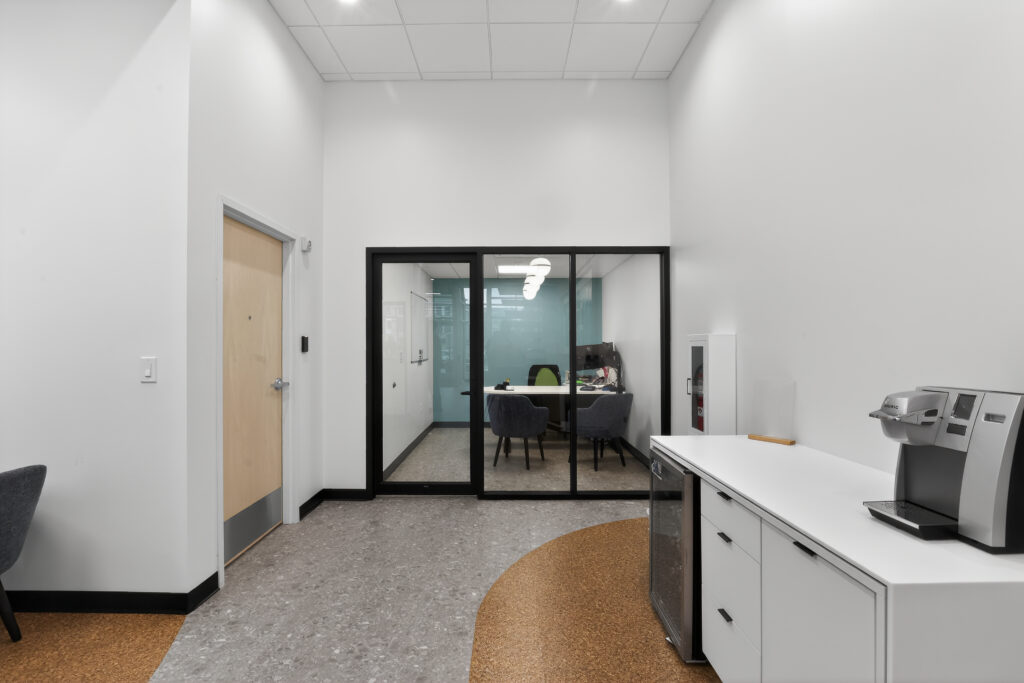 A small office with a glass wall and door, adjacent to a hallway containing a coffee machine on a white cabinet.