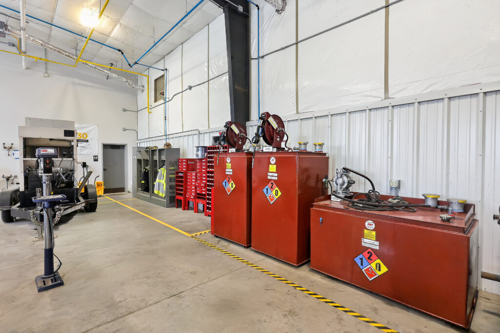 Three large red industrial fluid storage tanks with hazard labels in a warehouse, with a drill press and tool storage units in the background.