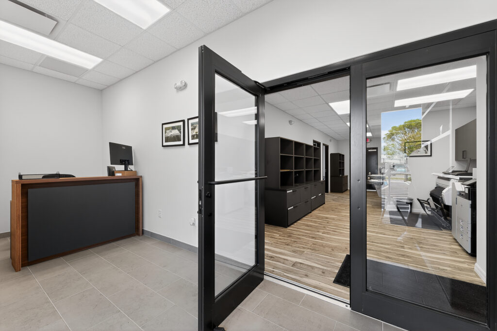 A modern office reception area with a wood and grey desk and an open glass door leading to a workspace with storage cabinets.