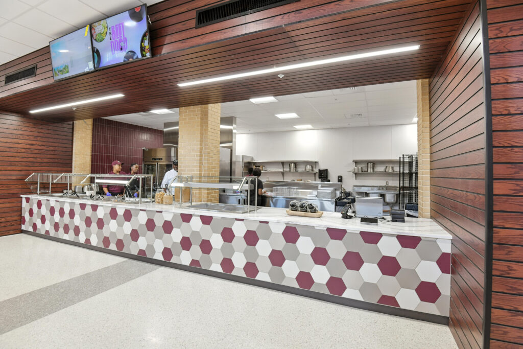 A cafeteria serving line with a geometric patterned counter, behind which staff work in a stainless-steel kitchen.