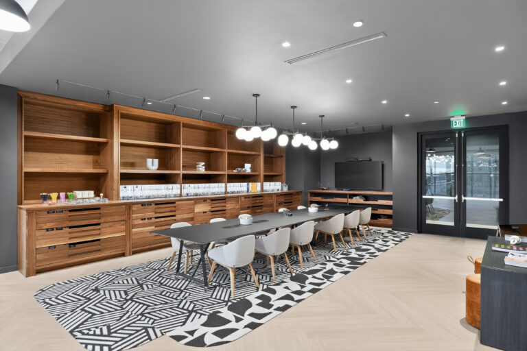 A long conference table with grey chairs sits on a geometric patterned rug in an office space with wooden storage walls and a television.