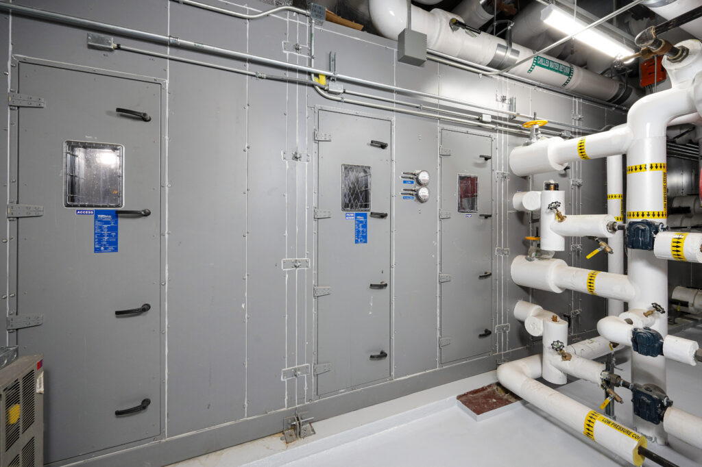 Industrial air handling unit with access doors and a series of white insulated pipes with flow labels.