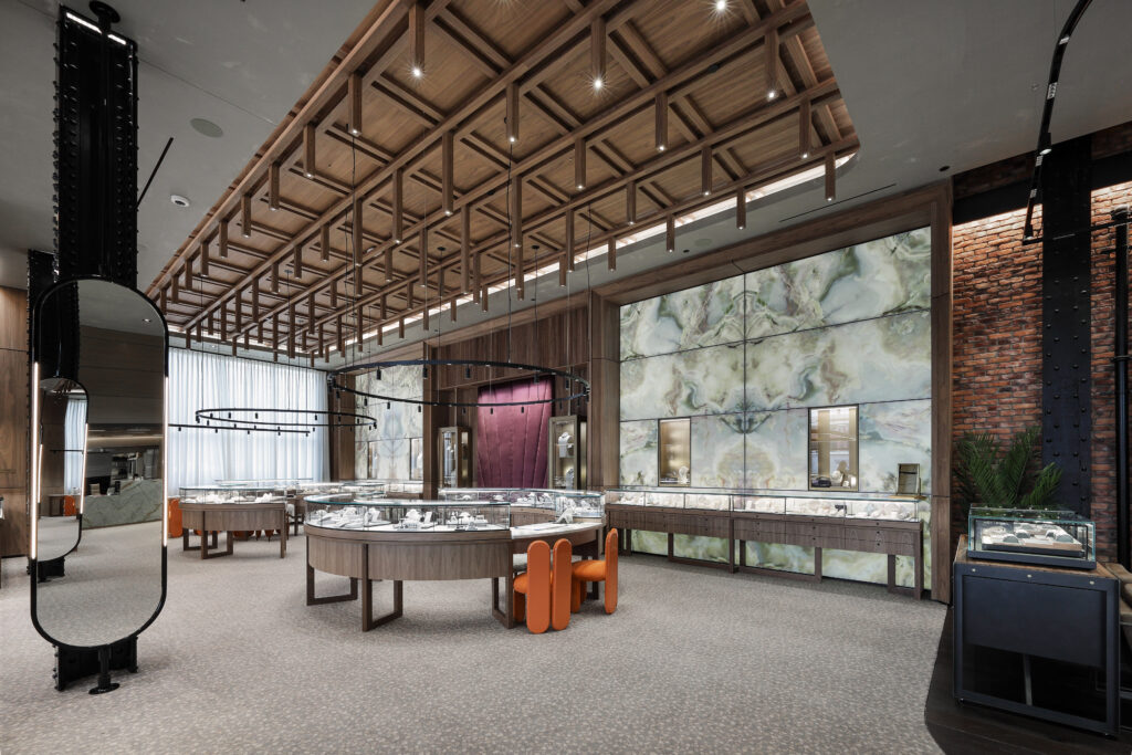 Interior of a jewelry store featuring glass display cases, orange chairs, a decorative coffered wooden ceiling, and a large backlit green stone feature wall.