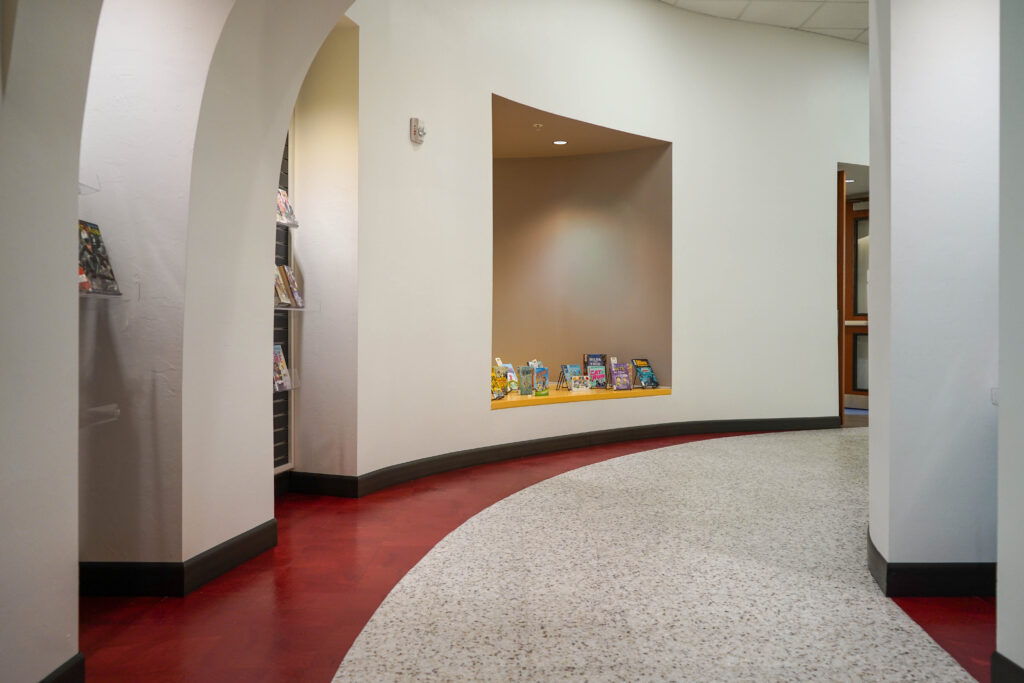 A curved hallway with red flooring and a central alcove displaying a row of books.