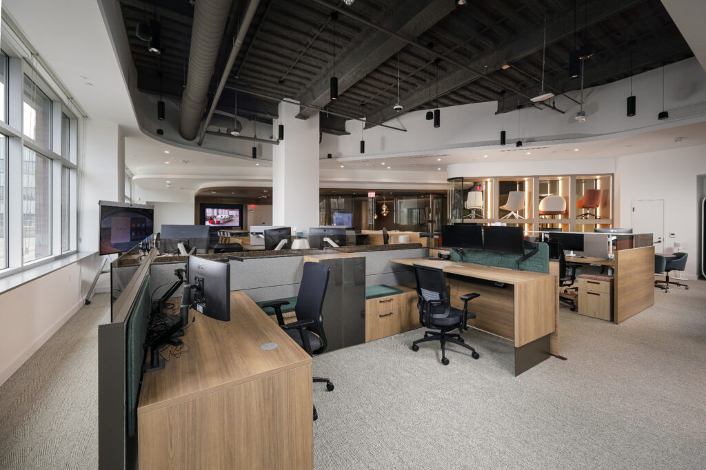 Modern open-plan office featuring rows of wooden desks with computers and ergonomic chairs, under an exposed ceiling with industrial ducts.