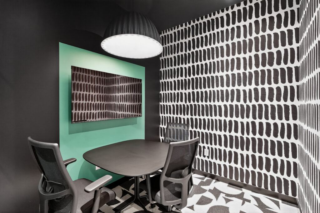 Small meeting room with a round table, three office chairs, a wall-mounted television, and patterned wallpaper.