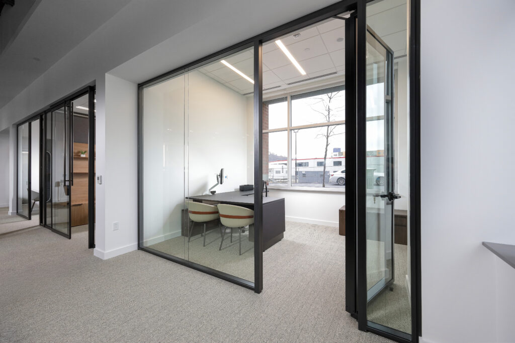 An interior view of a glass-walled office with a desk, two chairs, and a window looking out onto a parking lot.