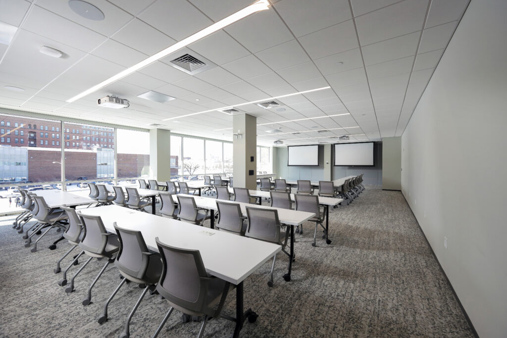 An empty conference room with rows of white tables, grey office chairs, and two projection screens.