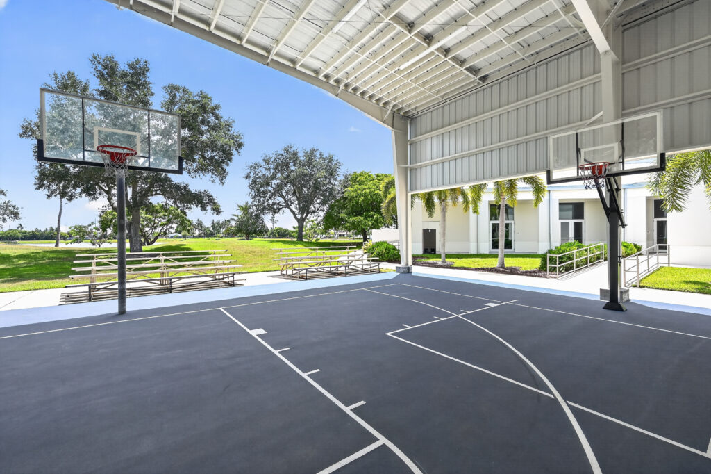 An outdoor basketball court under a large metal pavilion, with bleachers and trees in the background.