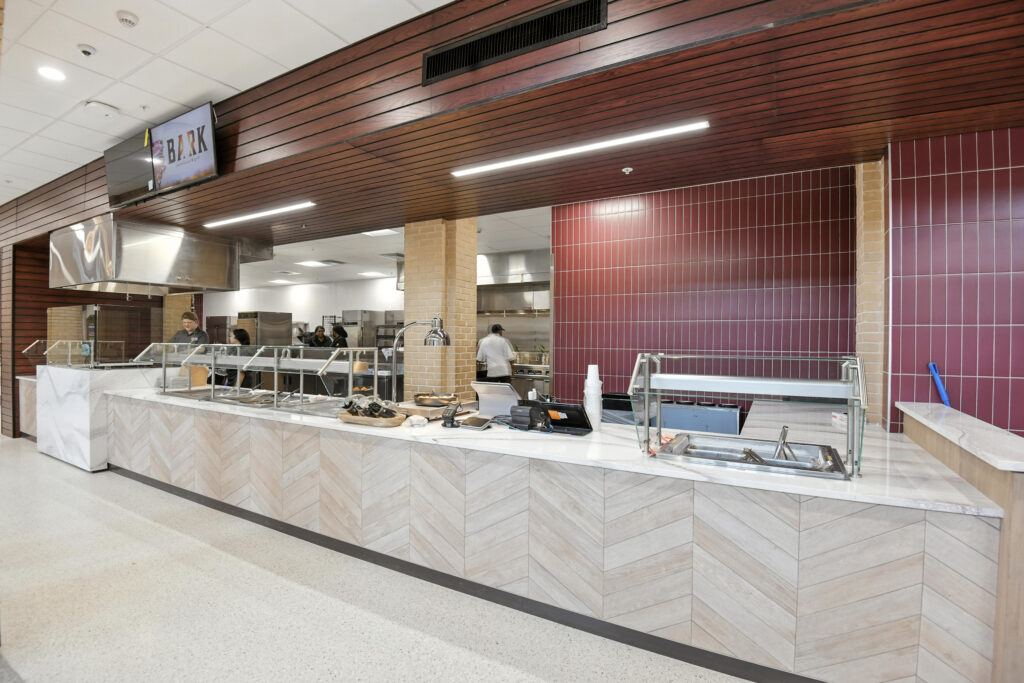 A cafeteria serving counter with a white herringbone pattern front, stainless steel sneeze guards, and a staff member visible in the background kitchen.