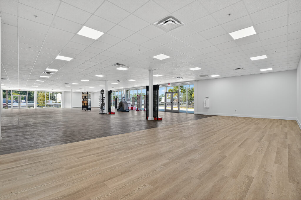 A large, empty retail space with light-toned wood-look flooring and floor-to-ceiling glass windows along the back wall.