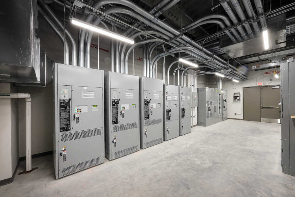 A row of industrial electrical switchgear cabinets in a utility room with exposed conduit piping on the ceiling.