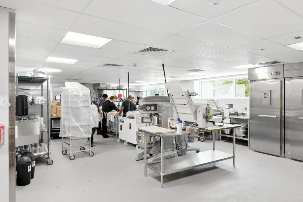Commercial kitchen with employees working at stainless steel prep tables, a large industrial mixer, and a Baxter oven.