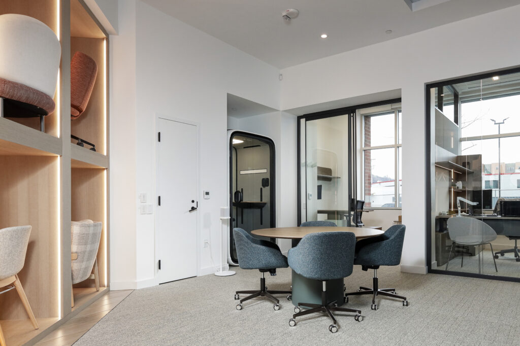 A small round meeting table with three blue swivel chairs in a modern office space, featuring a private phone booth and a glass-walled office in the background.