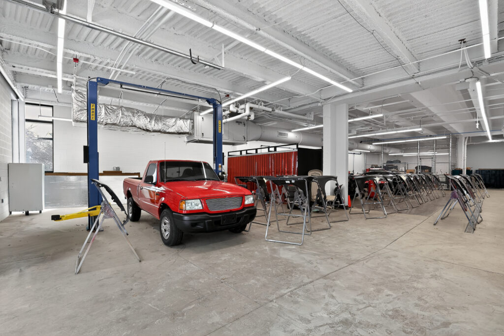 A red pickup truck parked in an auto body shop beside several car bumpers mounted on stands.