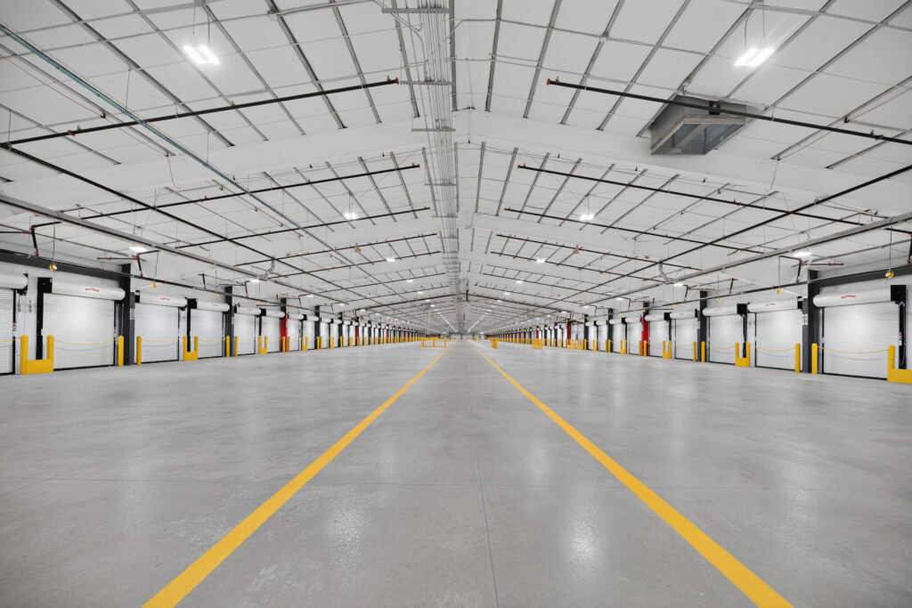 An empty industrial warehouse interior with two yellow floor markings leading to rows of closed loading dock doors on both sides.